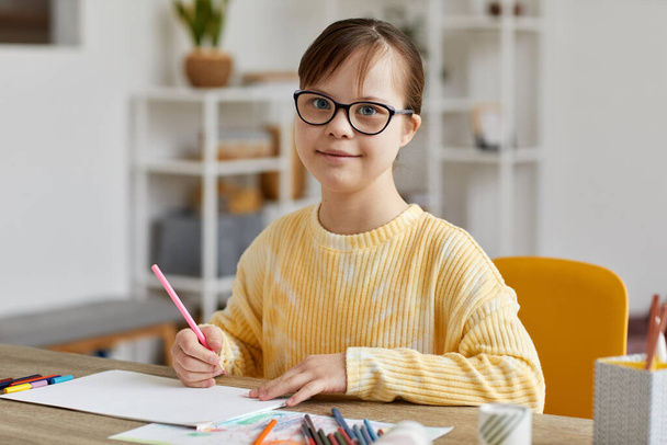 Portrait of cute teen girl with Down syndrome drawing pictures while sitting at desk and smiling at camera - Foto, Bild