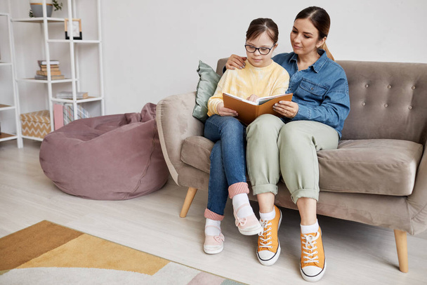 Full length portrait of teenage girl with Down syndrome reading book with loving mother at home - Foto, Bild