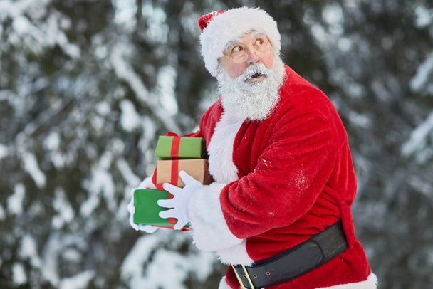 Retrato de la cintura hacia arriba de Papá Noel tradicional llevando regalos en secreto al aire libre y mirando cauteloso, espacio de copia - Foto, Imagen