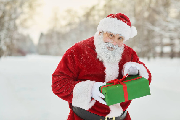 Waist up portrait of traditional Santa Claus holding gift box and looking at camera mischievously in winter forest, copy space - Photo, Image