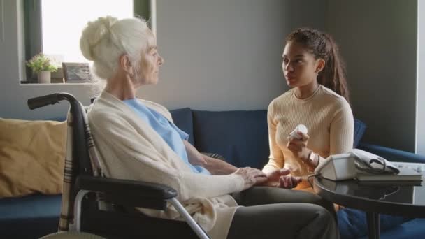 Молодая медсестра объясняет пожилой женщине в инвалидной коляске, как принимать лекарства и давать ей органайзер таблеток во время консультации по здоровью на дому - Кадры, видео