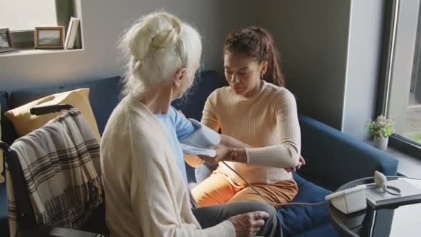 Mujer negra joven cuidadora midiendo la presión arterial de una mujer mayor sentada en silla de ruedas y hablando con ella durante el día en casa - Metraje, vídeo