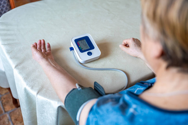 Idősebb nő ellenőrzi a vérnyomást otthon, idősebb nő szenved a magas vérnyomás, ül és használ egy pulzométer, tonométer. Visszanézni. Kiváló minőségű fénykép - Fotó, kép