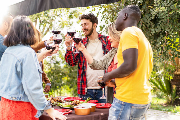 Ein lebhaftes Treffen unter freiem Himmel mit verschiedenen Freunden, die mit Weingläsern anstoßen und einen freudigen Anlass mit einem Grill im Hintergrund feiern. - Foto, Bild