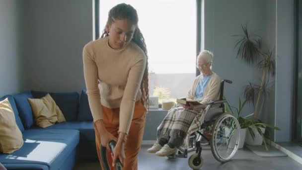 Gekippte Aufnahme einer jungen Pflegerin beim Staubsaugen des Teppichs im Wohnzimmer, während sie einer älteren Frau im Rollstuhl bei der Hausreinigung hilft - Filmmaterial, Video