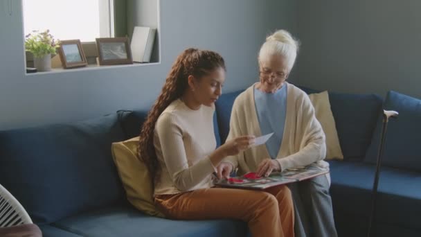 Pflegerin sitzt auf Sofa im Wohnzimmer, schaut Fotoalbum und unterhält sich mit älterer Frau, während sie sie zu Hause besucht - Filmmaterial, Video