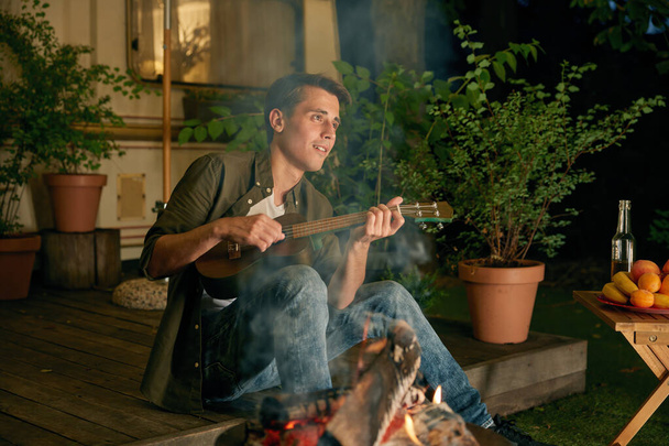 Jovem tocando guitarra e cantando música romântica na fogueira do trailer. Homem casual, estilo de vida e férias de verão conceito ao ar livre - Foto, Imagem