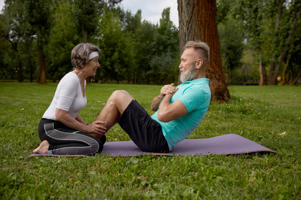 Ευτυχισμένο παντρεμένο ζευγάρι συνταξιούχων που κάνουν προπόνηση στο πάρκο. Ηλικιωμένη σύζυγος που βοηθάει τον ηλικιωμένο σύζυγο να κάνει κοιλιακή κρίση. Αθλητική δραστηριότητα αναψυχής για συνταξιούχους - Φωτογραφία, εικόνα