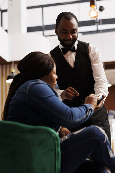 Afroameričanka host bezkontaktní platby v hotelu, zatímco odpočívá v hale. Usmívající se přátelský číšník pomocí čtečky karet NFC umožňuje zákazníkovi zaplatit za objednávku v restauraci nebo kavárně - Fotografie, Obrázek