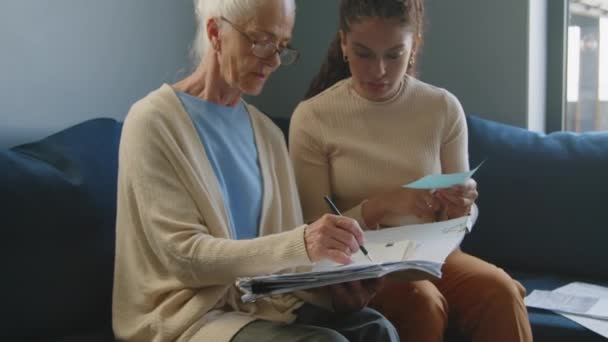 Νεαρή γυναίκα φροντιστής διαβάζοντας λογαριασμούς και μιλώντας με ηλικιωμένη γυναίκα κρατώντας σημειώσεις στο προσωπικό οικονομικό βιβλίο εργασίας, ενώ το σχεδιασμό του προϋπολογισμού μαζί στο σπίτι - Πλάνα, βίντεο