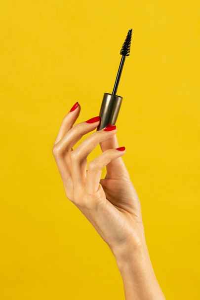 Anonymous young slim female with nails polished slender fingers holding aloft mascara base gently and demonstrating against yellow background in light - Photo, Image