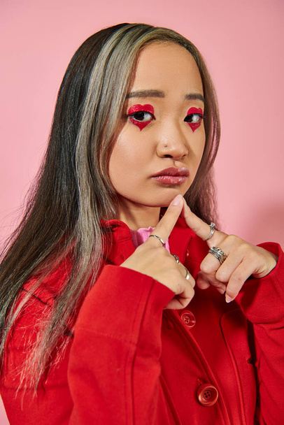 pensativo joven asiático mujer con corazón en forma de ojo maquillaje y teñido pelo mirando cámara en rosa - Foto, Imagen