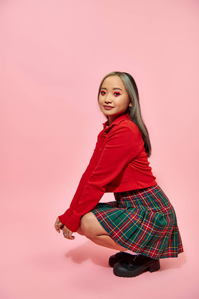 pleine longueur de jeune femme asiatique avec coeur en forme de maquillage des yeux et jupe à carreaux assis sur rose - Photo, image