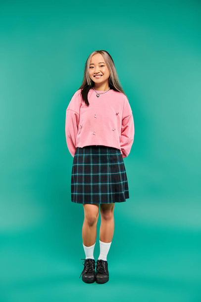 full length of cheerful asian woman in pink sweatshirt and plaid skirt standing on blue backdrop - Photo, Image
