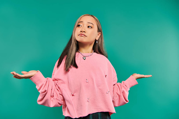 portrait of confused asian woman in pink sweatshirt and heart shaped necklace on blue backdrop - Photo, Image