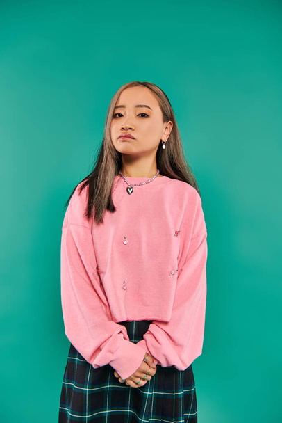 portrait of pensive asian woman in pink sweatshirt and plaid skirt standing on blue backdrop - Photo, Image
