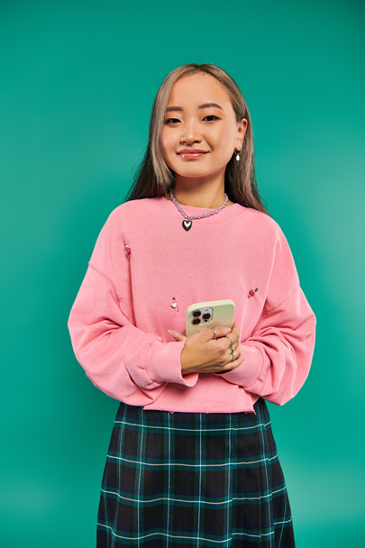 портрет счастливой молодой азиатки в розовой толстовке и клетчатой юбке с помощью смартфона на бирюзе - Фото, изображение