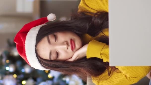 Vídeo vertical de menina asiática bonita vestindo chapéu de Papai Noel derrubando triste enquanto sentado em casa no sofá com laptop em seu colo. Mulher bonita celebrando o Natal sozinha no quarto aconchegante. Feriado solitário. - Filmagem, Vídeo