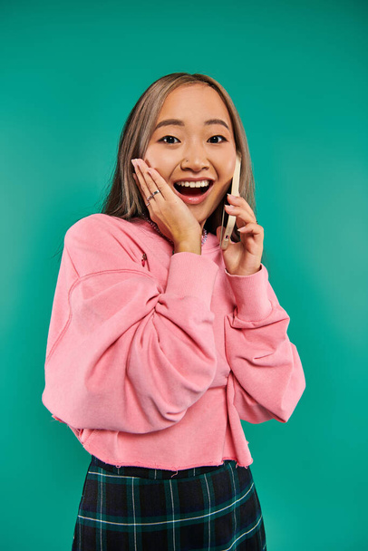 портрет изумленной молодой азиатки в розовой толстовке разговаривающей по смартфону на бирюзовом фоне - Фото, изображение