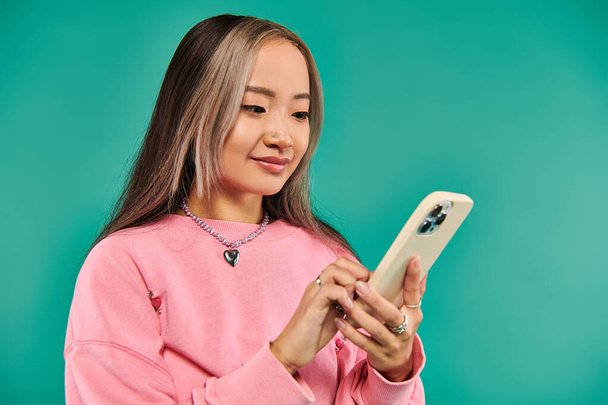 retrato de la joven mujer asiática complacida en sudadera rosa y falda a cuadros usando teléfono inteligente en azul - Foto, Imagen