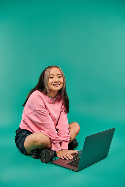 szczęśliwy Azji kobieta w bluzie i spódnica w kratę siedzi i za pomocą laptopa na turkusowym tle - Zdjęcie, obraz
