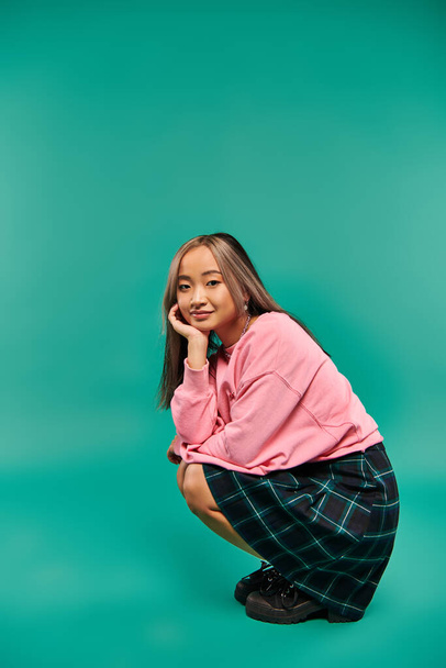 full length of pleased young asian woman in pink sweatshirt and plaid skirt sitting on pink backdrop - Photo, Image