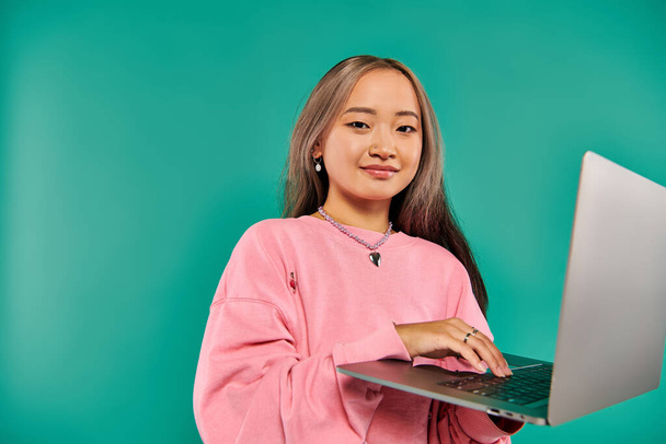 задоволена азіатська жінка в рожевому светрі, використовуючи ноутбук, стоячи на бірюзовому фоні, мережа - Фото, зображення