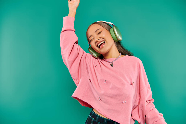 positive junge asiatische Mädchen in drahtlosen Kopfhörern gestikulieren und tanzen auf türkisfarbenem Hintergrund - Foto, Bild