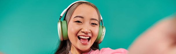 χαρούμενο νεαρό κορίτσι από την Ασία με ασύρματα ακουστικά που ακούει μουσική και βγάζει selfie σε τυρκουάζ, banner - Φωτογραφία, εικόνα