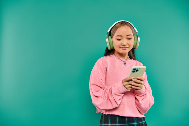 heureux asiatique fille dans sans fil casque écoute musique et en utilisant smartphone sur turquoise toile de fond - Photo, image
