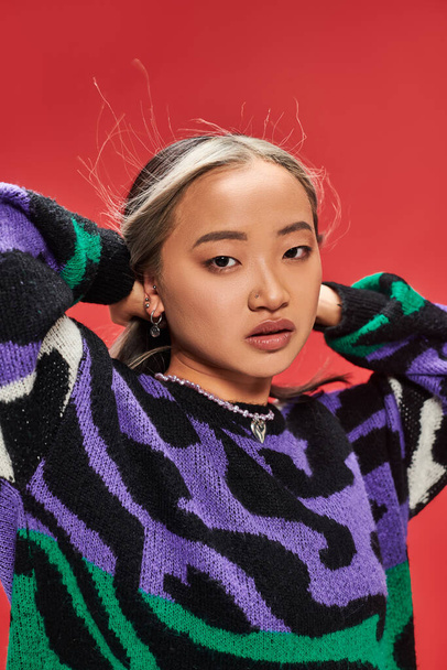 charming young asian woman in vibrant sweater with animal print adjusting hair on red backdrop - Photo, Image