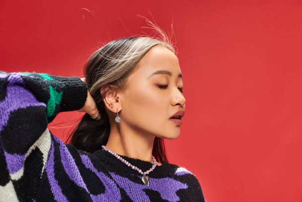 赤い背景に髪を調整する動物プリントと活気のあるセーターで魅力的な若いアジアの女性 - 写真・画像