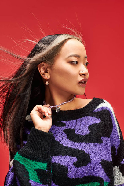 charming young asian woman in vibrant sweater with animal print touching necklace on red backdrop - Photo, Image