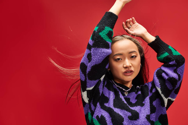 pretty young asian woman in vibrant sweater with animal print posing with raised hands on red - Photo, Image