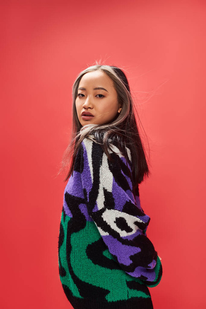 junges asiatisches Model in lebendigem Pullover mit Animal Print posiert auf rotem Hintergrund, Blick in die Kamera - Foto, Bild