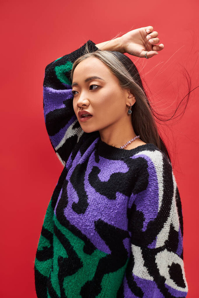 beautiful and young asian woman in knitted sweater with animal print posing on red background - Zdjęcie, obraz