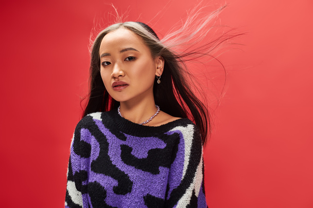 wind in hair of beautiful young asian woman in sweater with animal print posing on red background - Photo, Image