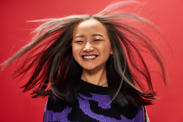 vent dans les cheveux de joyeuse jeune femme asiatique en pull avec imprimé animal posant sur fond rouge - Photo, image