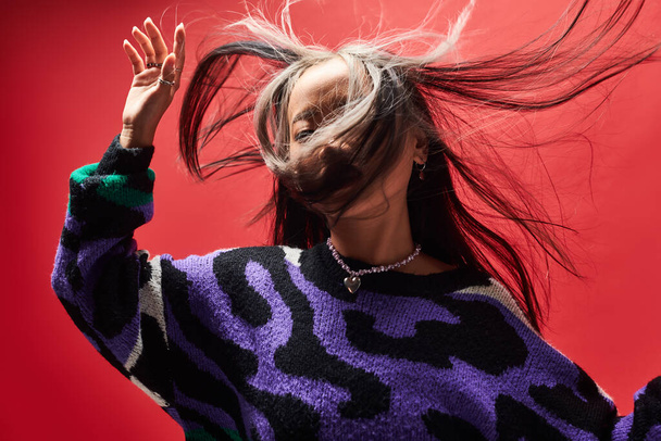 wind in haar van jong aziatisch model in trui met dierenprint gebaren op rode achtergrond - Foto, afbeelding
