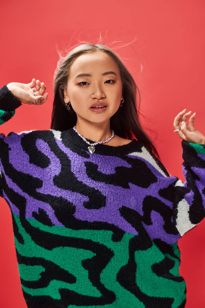 young asian girl in sweater with animal print with heart shaped necklace posing on red backdrop - Photo, Image