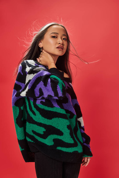 young asian woman in sweater with animal print touching neck while posing on red backdrop - Photo, Image