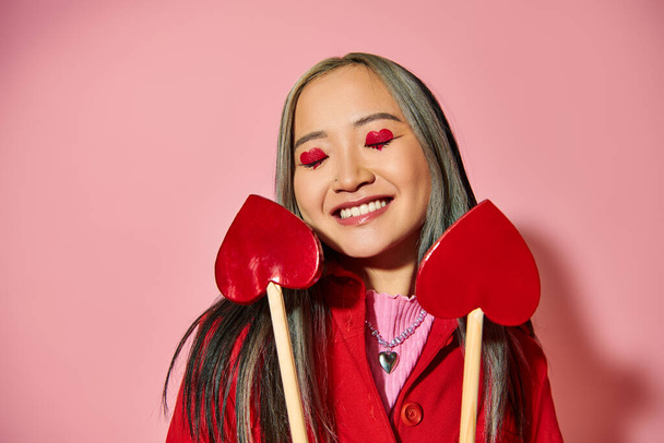 Valentines day concept, jolly asian girl with heart shaped eye makeup holding arrows on pink - Photo, Image