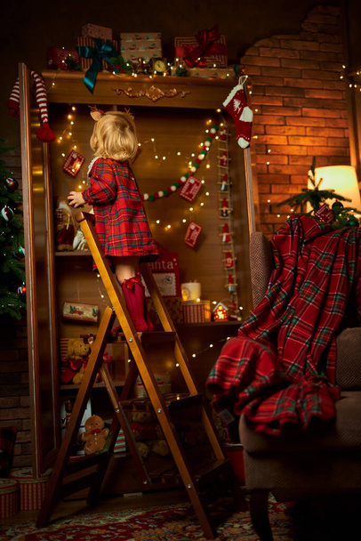Joyeux Noël et Joyeuses Fêtes. Une petite fille en robe à carreaux monte les escaliers jusqu'à l'étagère supérieure d'un placard avec des cadeaux. Atmosphère de magie, d'enchantement et de célébration - Photo, image