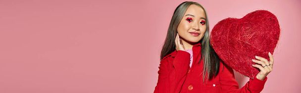 Valentines day, happy asian woman with vibrant eye makeup holding red heart on pink backdrop, banner - Photo, image