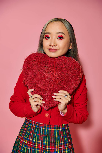 San Valentín día, positivo asiático mujer con vibrante ojo maquillaje celebración rojo corazón en rosa telón de fondo - Foto, Imagen