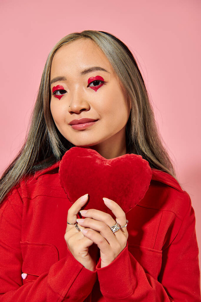 Valentines day, cheerful asian woman with vibrant eye makeup holding red heart on pink backdrop - Photo, Image