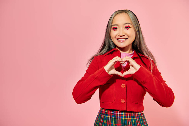 Ημέρα του Αγίου Βαλεντίνου, ευτυχισμένη ασιατική κοπέλα με κόκκινο μακιγιάζ ματιών δείχνει την καρδιά με τα χέρια σε ροζ φόντο - Φωτογραφία, εικόνα