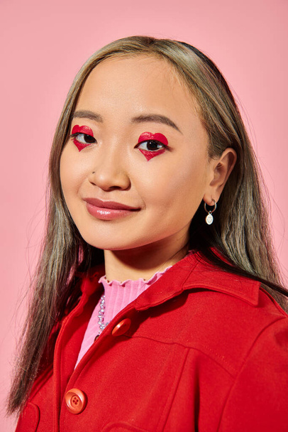 День Святого Валентина, усміхнена азіатська молода жінка з макіяжем для очей позує в червоній куртці на рожевому - Фото, зображення