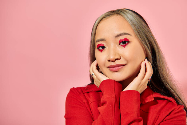 Valentim dia conceito, bonito asiático jovem mulher com coração olho maquiagem posando no vermelho jaqueta no rosa - Foto, Imagem