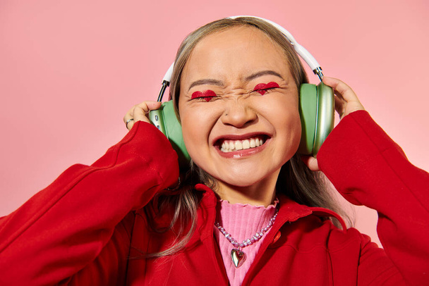 radostná mladá asijská žena v zelených bezdrátových sluchátkách s make-upem ve tvaru srdce na růžovém pozadí - Fotografie, Obrázek